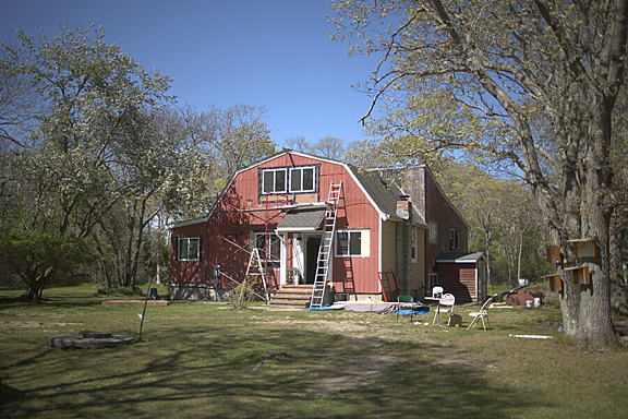 A red house under construction with ladders leaning against the front stands surrounded by trees