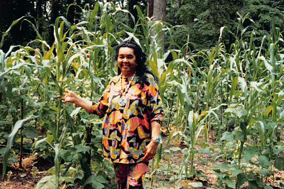 A women in bright cloths stands smiling in front of rows of tall plants