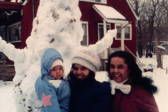 Two women smile holding a child stand outside in front of a snow man and a red house
