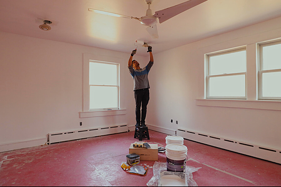 A man stands on a small step stool in an empty room, painting the ceiling with a paint roller