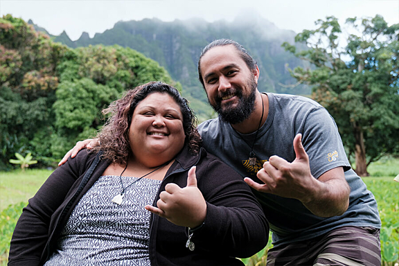 A man and women smile at camera while outside making a the Mahalo hand gesture