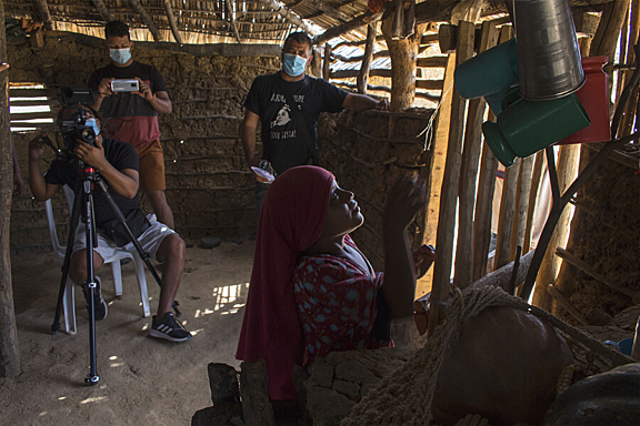 Two men film a women while she sits in front of a group of bottles