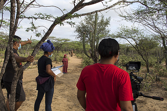 A man in red shirt films a women in the background while two people watch over