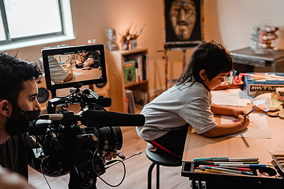A child colors at his desk while being filmed by a camera operator.