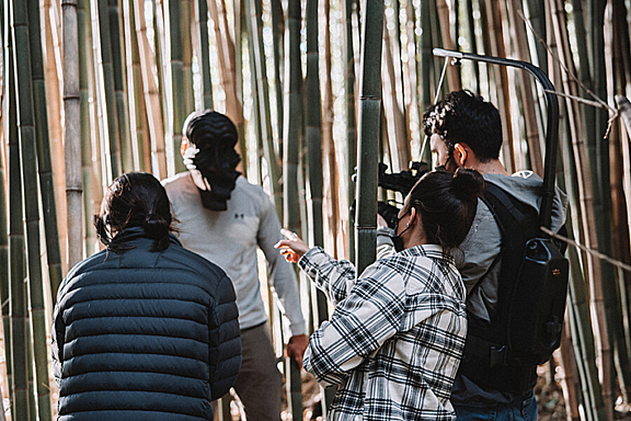 A women points in a group with a camera operator and a man in carved wooded mask