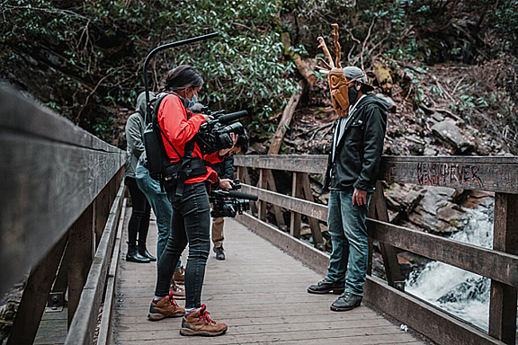 A woman in red jacket films a man in a carved wooden mask on a bridge