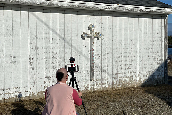 A person in a pink vest kneels to take a photo in front of a white building with a cross