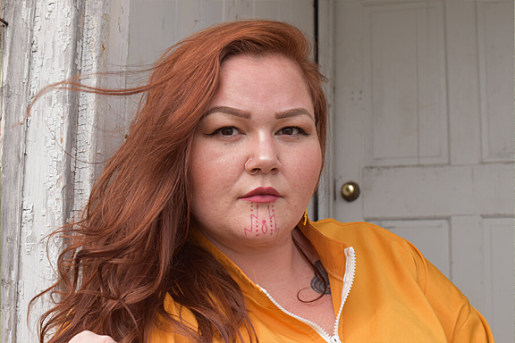 A women with red hair and small chin tattoo stares intently at camera