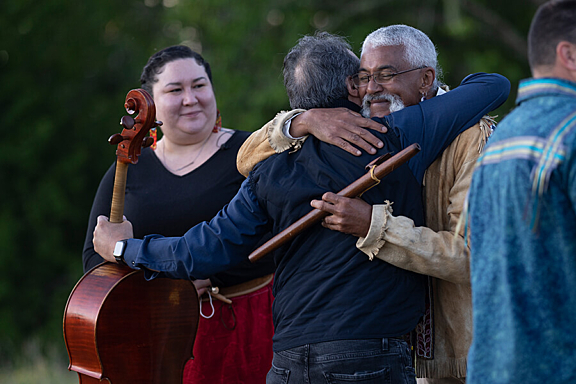 A man with a cello hugs another man with a flute while a woman smiles behind them