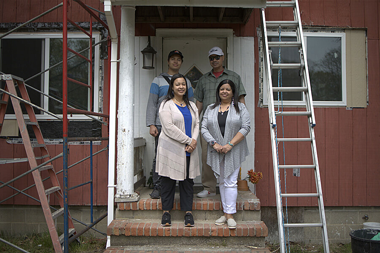 Two men and two women stand on the stoop of a red house smiling surrounded by ladders and scaffolding