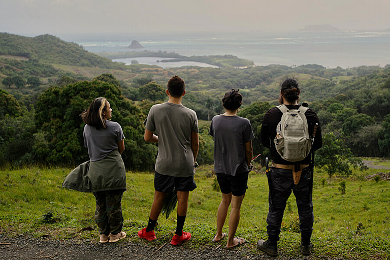 Four people stand on a hillside looking outward over threes and water