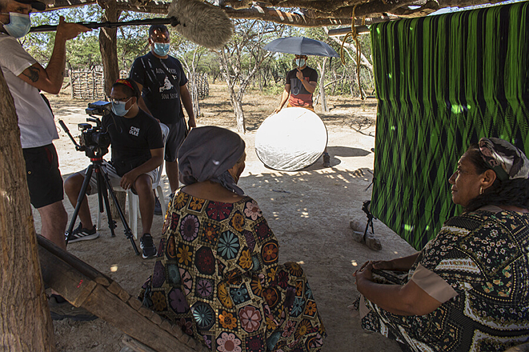 Two women in traditional patterned dresses sit and talk while four men with film equipment record behind them