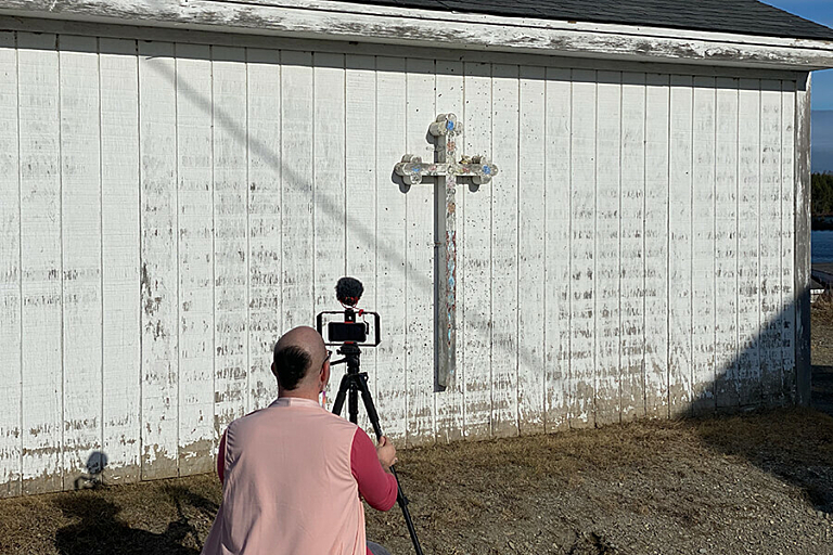 A person in a pink vest kneels to take a photo in front of a white building with a cross
