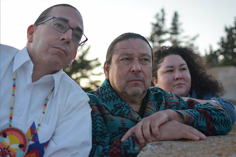 A man in glasses leans on a rock next two another man and women looking in the distance