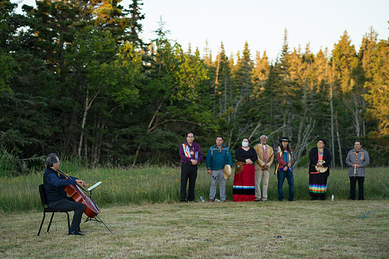 A man plays a cello outdoors while a group of seven people watch from a distance