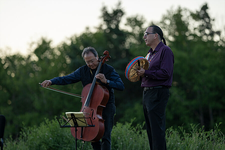 A man plays a cello and another man plays a hand drum while standing in a forest at dawn